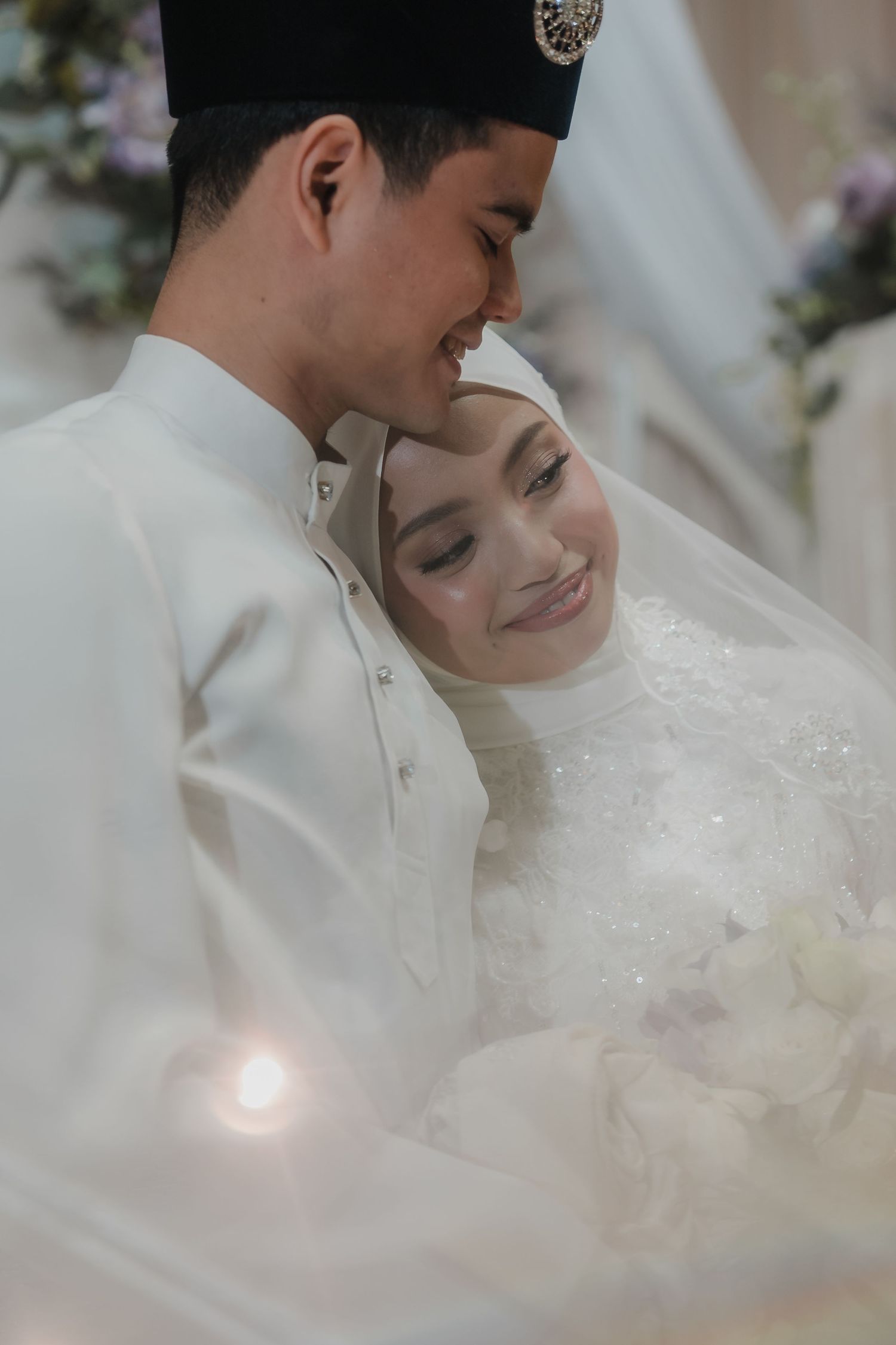 A Muslim couple in white wedding attire share a tender moment, the bride smiling in hijab as the groom leans close.