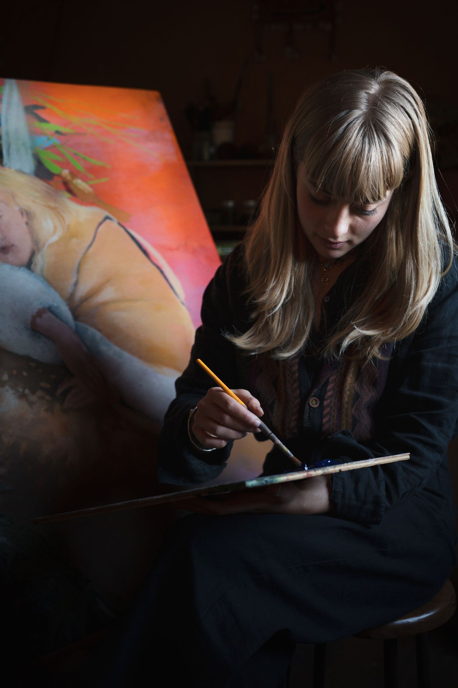 Blonde female artist sitting and painting on canvas with brush and palette in dark moody studio.

Maya Caroll-Tischler