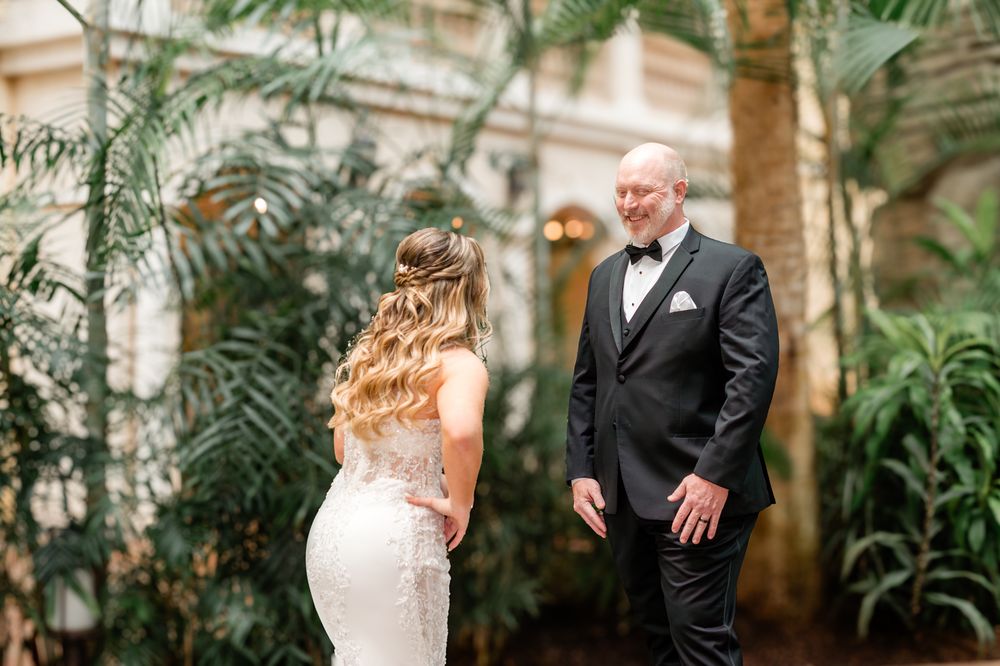 Bride in fitted lace gown shares first look moment with man in black tuxedo among tropical plants.