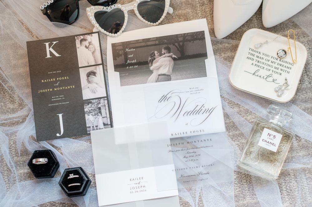 Elegant wedding stationery flat lay with invitations, rings, perfume, and heart sunglasses on lace fabric.