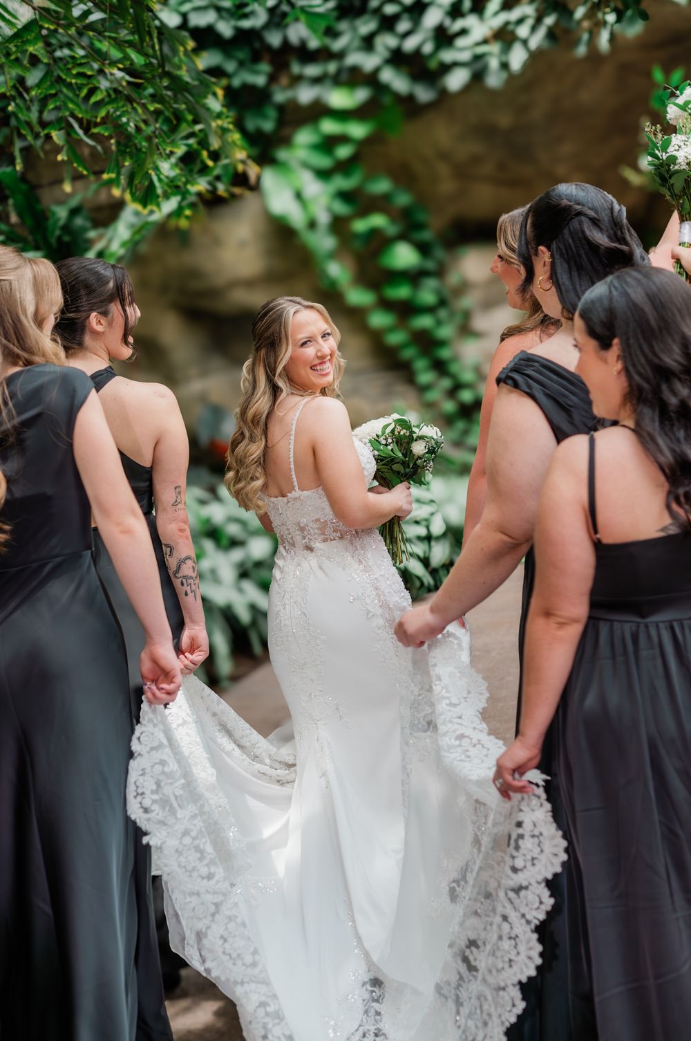 Bride in lace wedding gown smiles at bridesmaids in black dresses holding bouquets in garden setting.