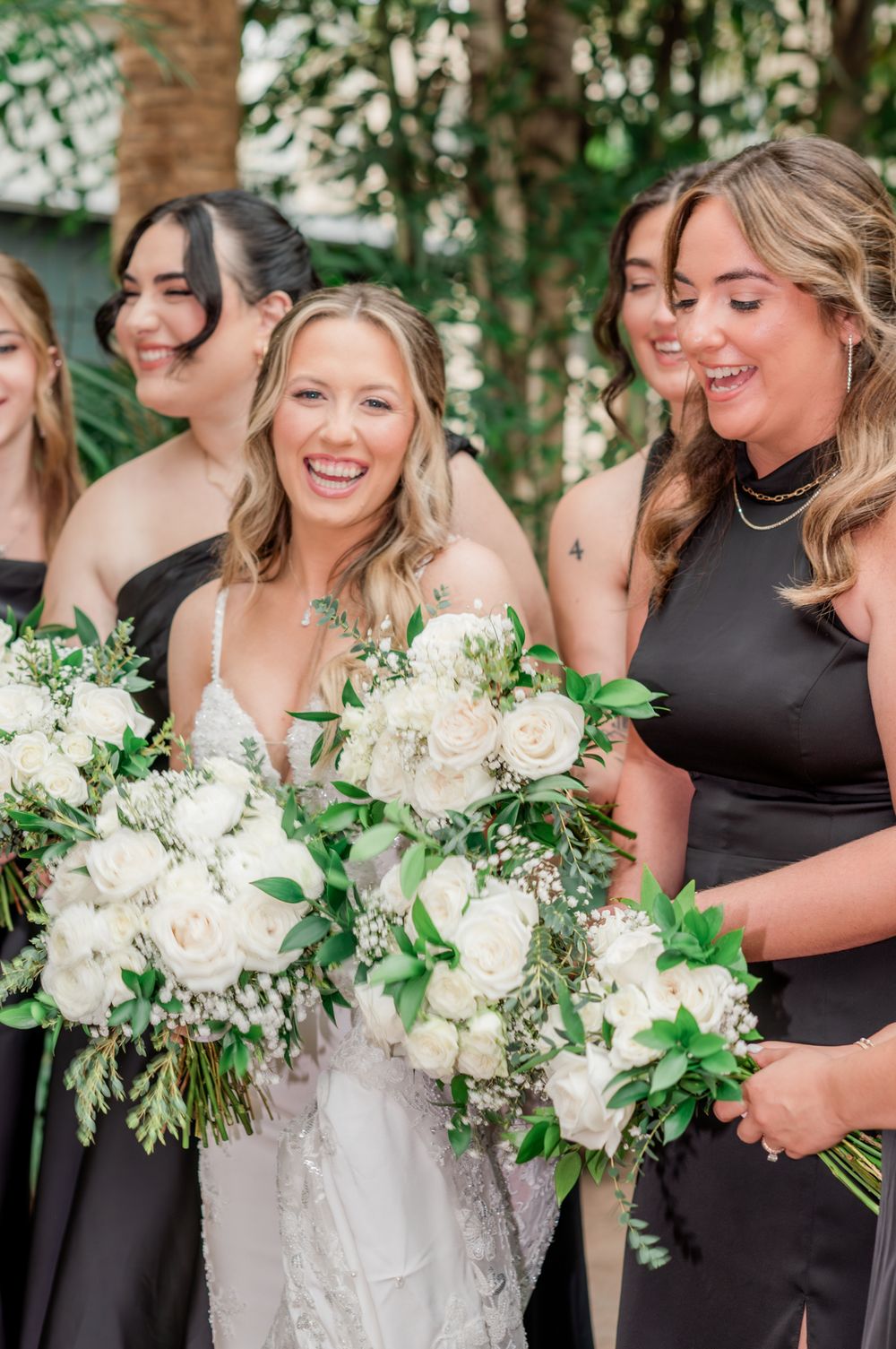 Bride laughing with bridesmaids in black dresses holding white rose and greenery bouquets outdoors.