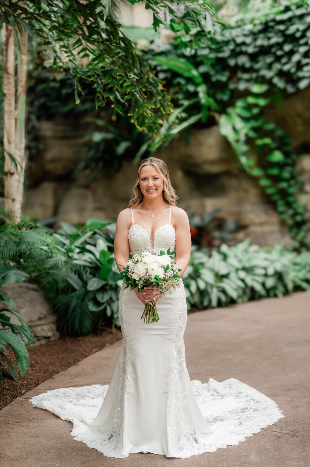 Bride in fitted lace wedding gown holds white bouquet while posing in a lush garden setting.