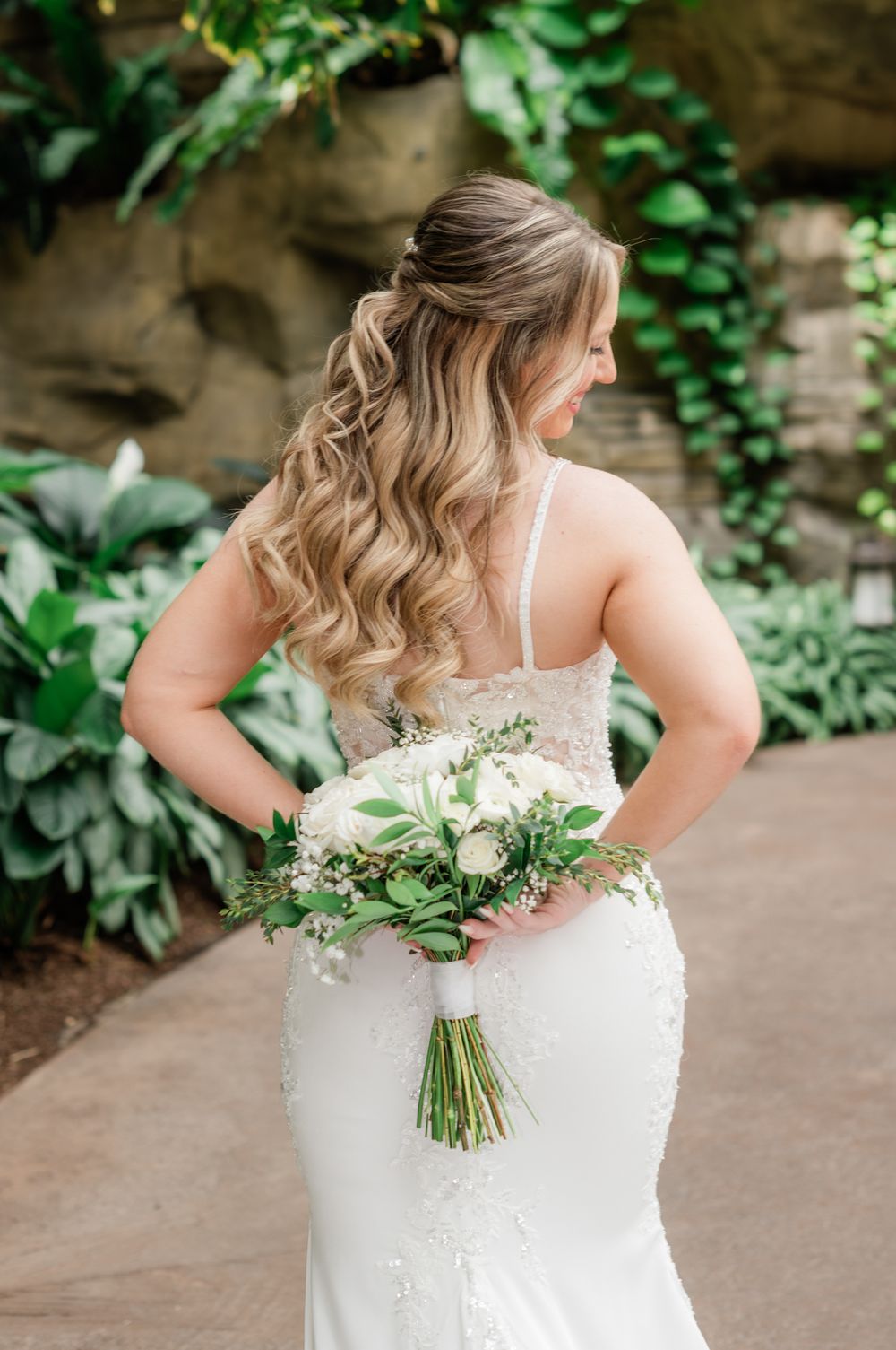 Bride in fitted lace wedding gown holds white floral bouquet with greenery, showing off her wavy blonde hair.