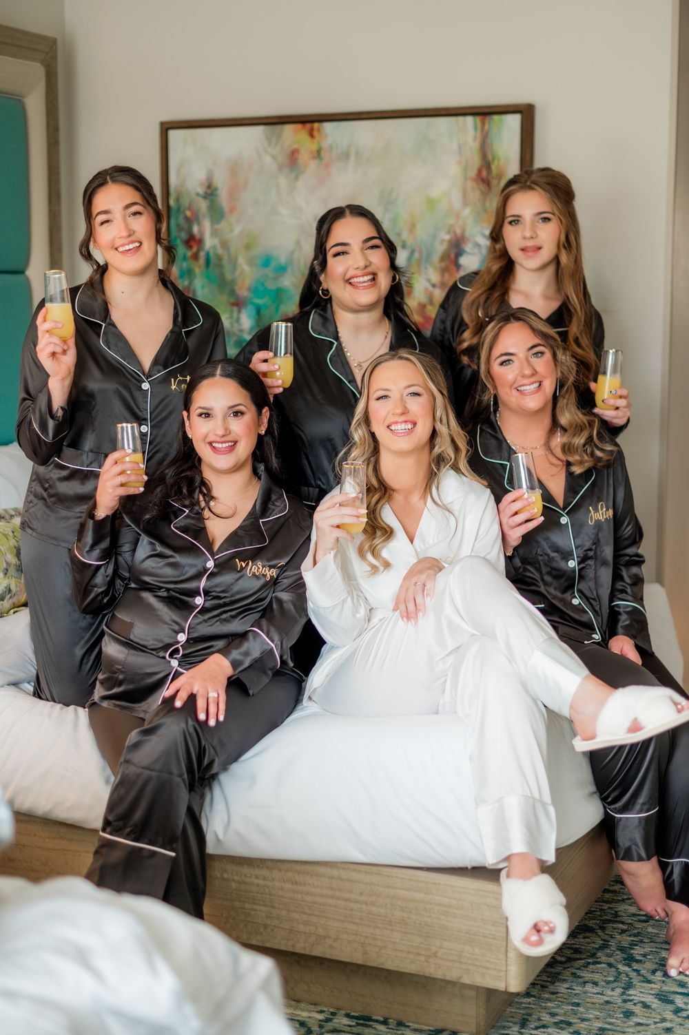 Bride in white satin robe poses with bridesmaids in black pajamas holding champagne flutes on a bed.