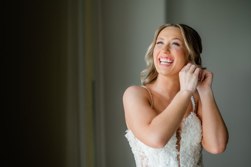 Smiling bride in lace wedding dress adjusting earring near window in soft natural light.