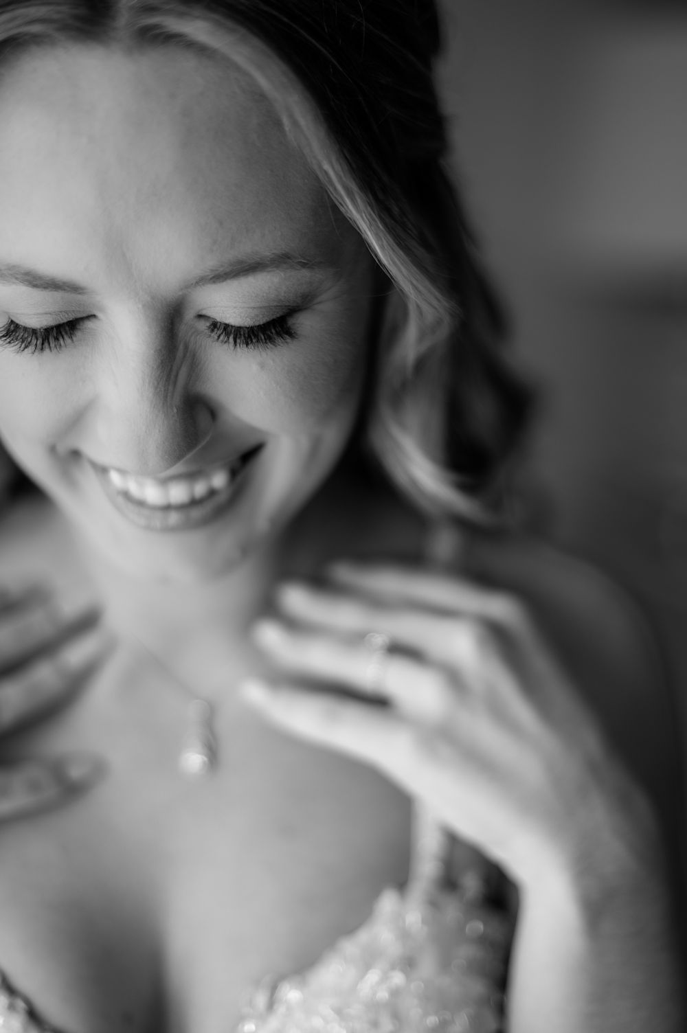 Black and white bridal portrait of smiling bride with pearl necklace and engagement ring, eyes closed, lace dress.