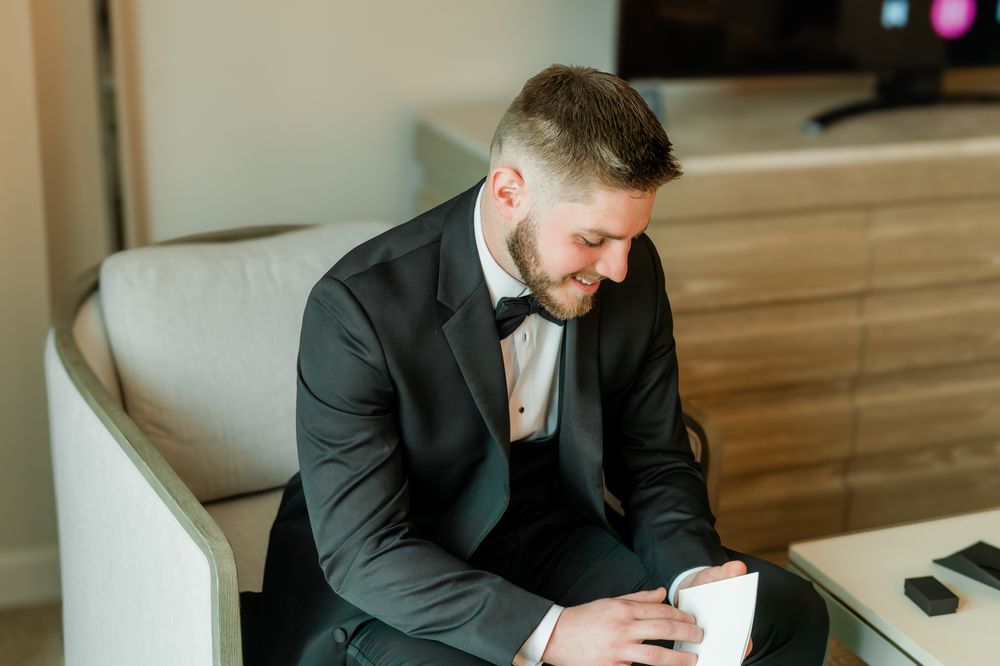 Groom in black tuxedo with bow tie reading a letter while seated in a chair on his wedding day.