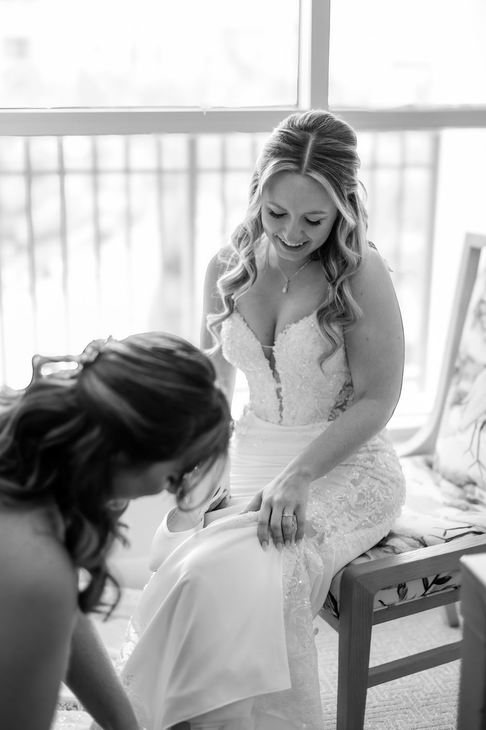 Bride in lace wedding gown smiles as bridesmaid helps with dress preparations near a bright window.