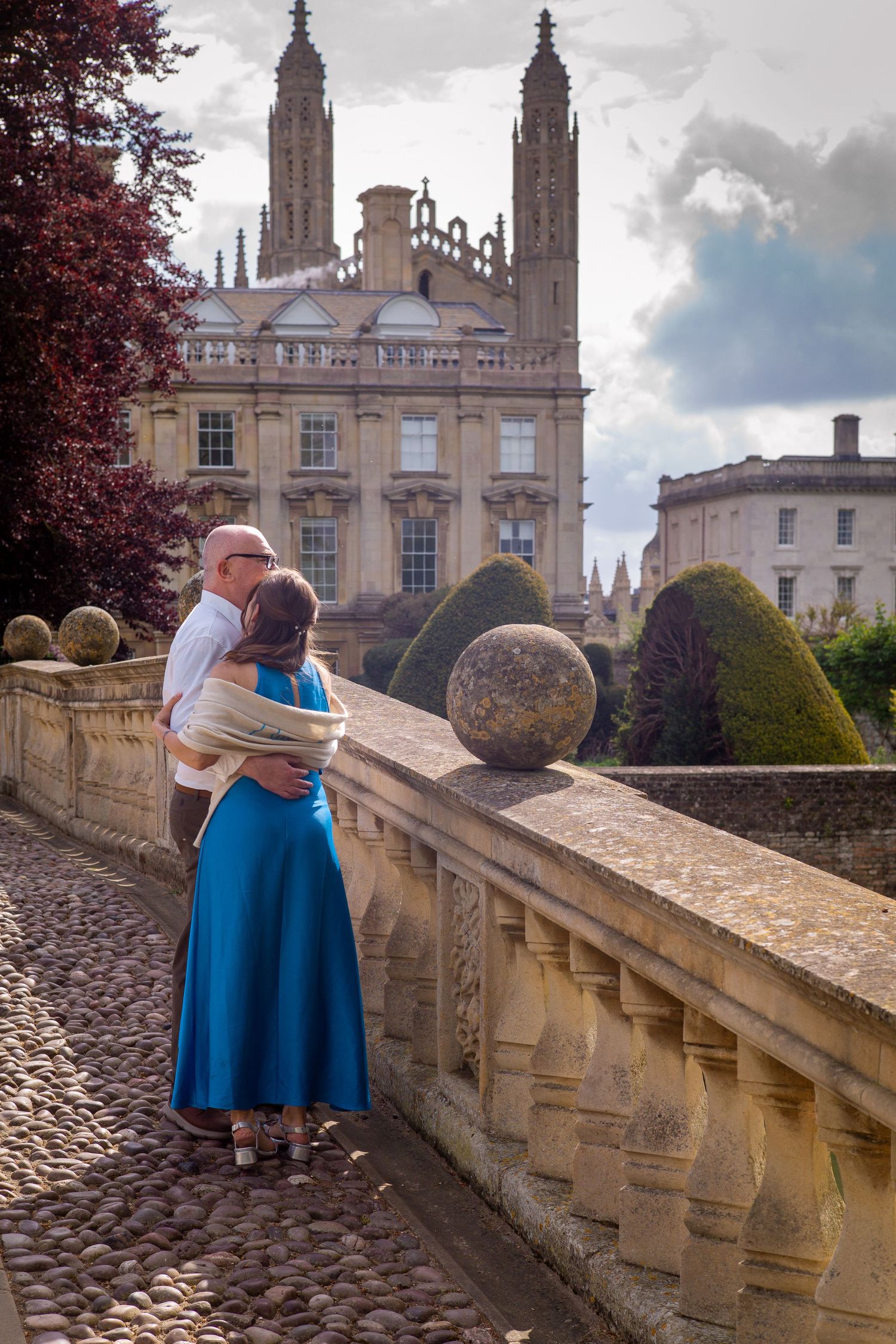 Couple sitting on stone steps, man in dark jacket embracing woman in blue skirt, outdoor urban setting.