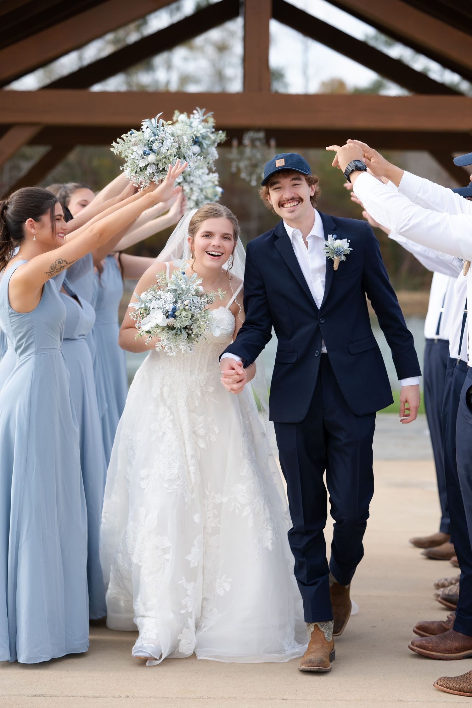 Bride and groom walk through arch of raised bouquets with bridesmaids in blue gowns and groomsmen at outdoor wedding.