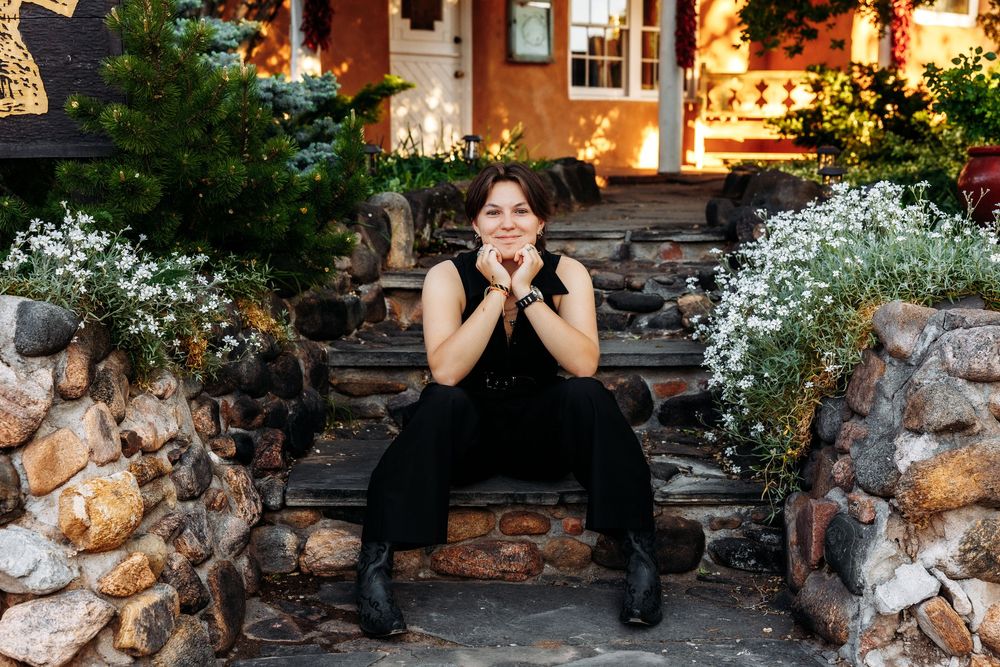 Woman in black outfit sitting on stone steps surrounded by white flowers and greenery outside a warmly lit building.