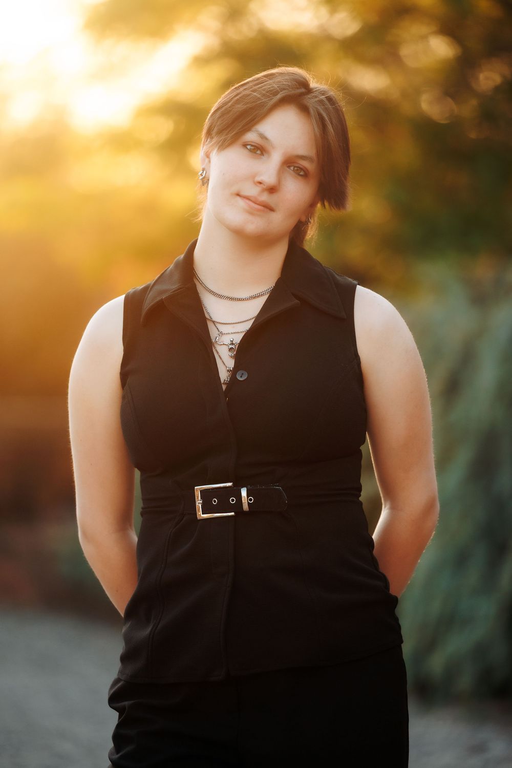 Young woman in black sleeveless dress with belt poses outdoors during golden hour sunset.