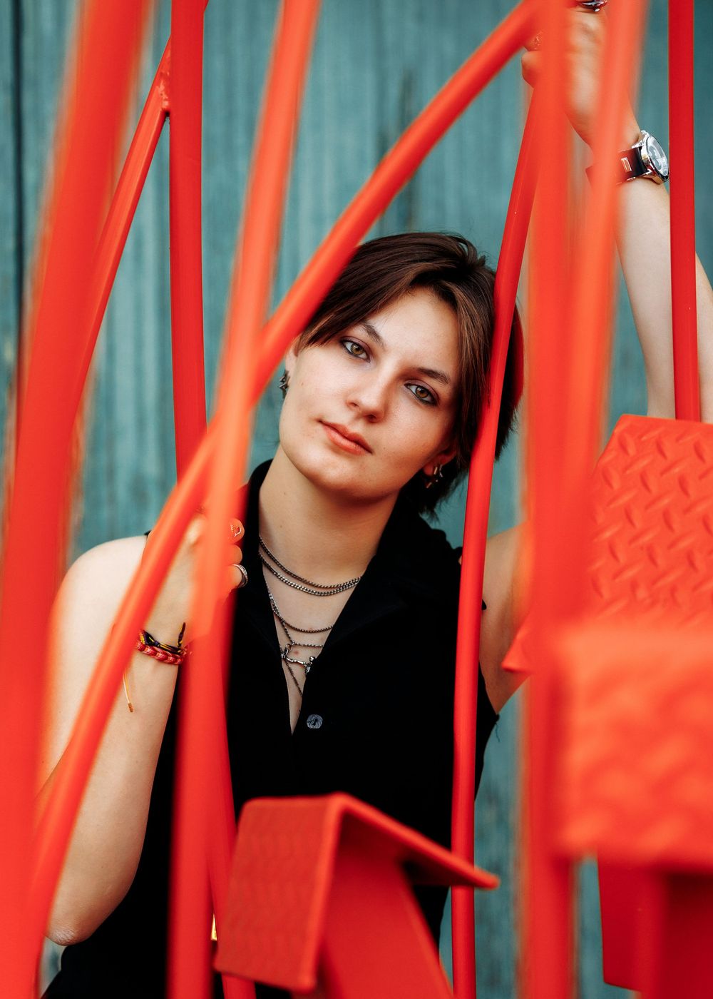 Young woman with dark hair posing among vibrant red metal structures against a teal wooden background.