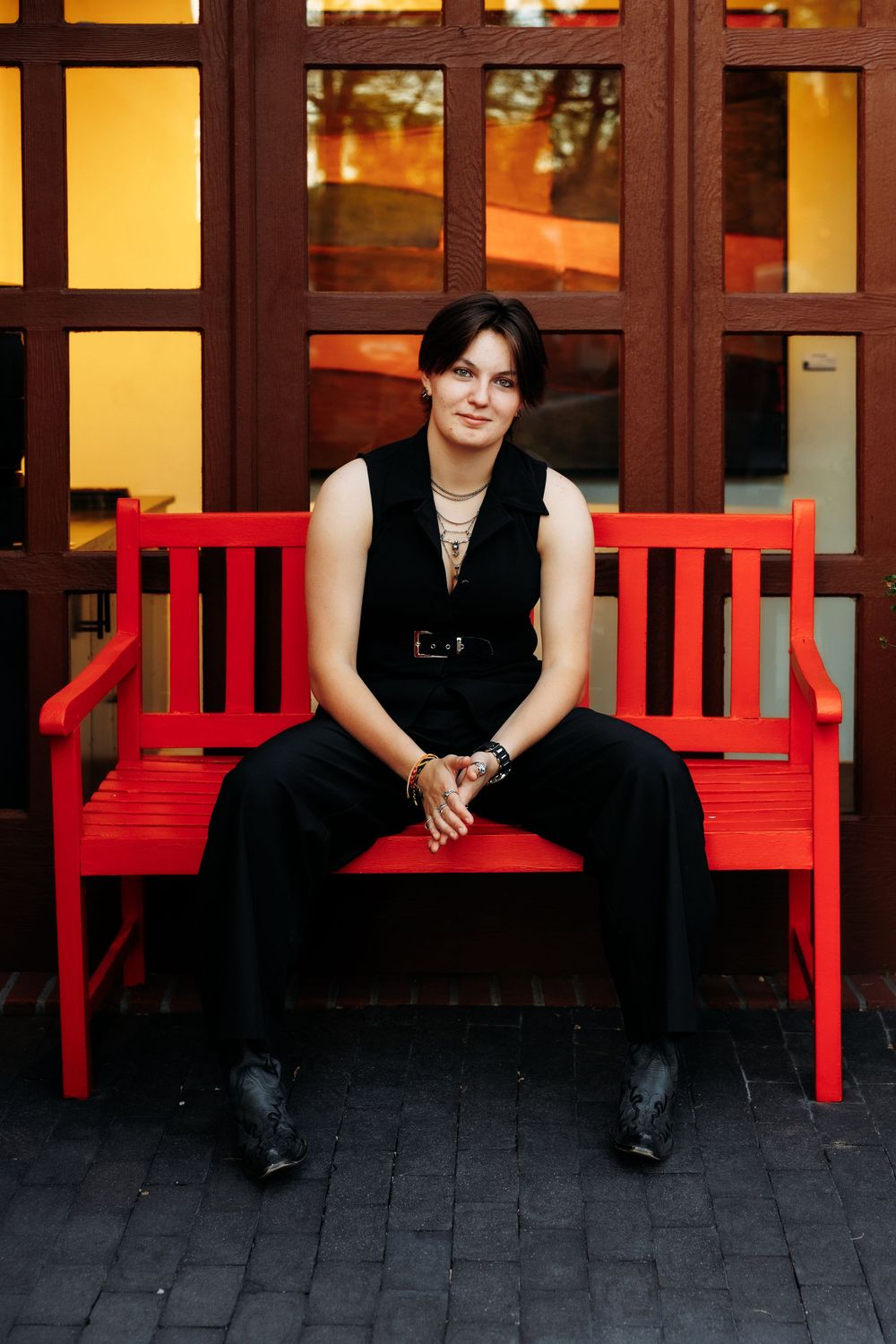 A young woman in black outfit sits on a vibrant red wooden bench in front of a building with large windows.