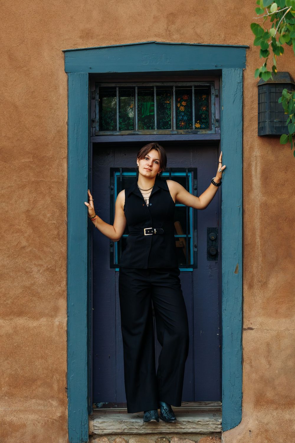 Woman in black outfit posing in a blue-framed doorway of an adobe building.