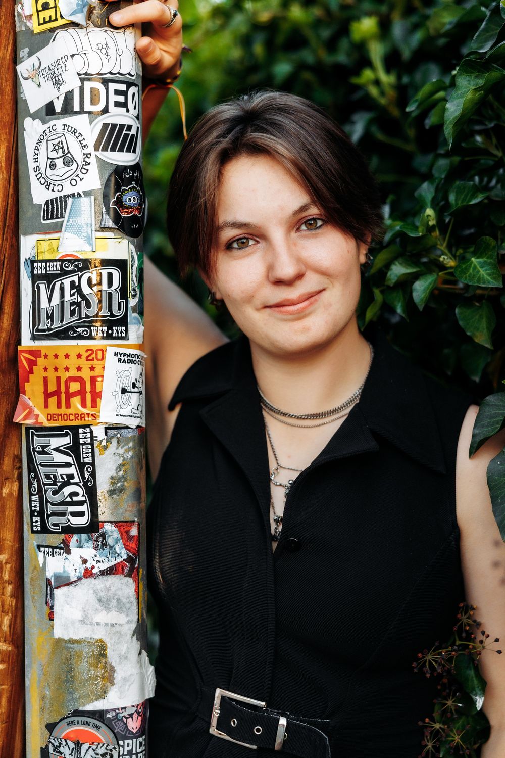 Young woman in black outfit smiling beside a sticker-covered pole with green foliage background.