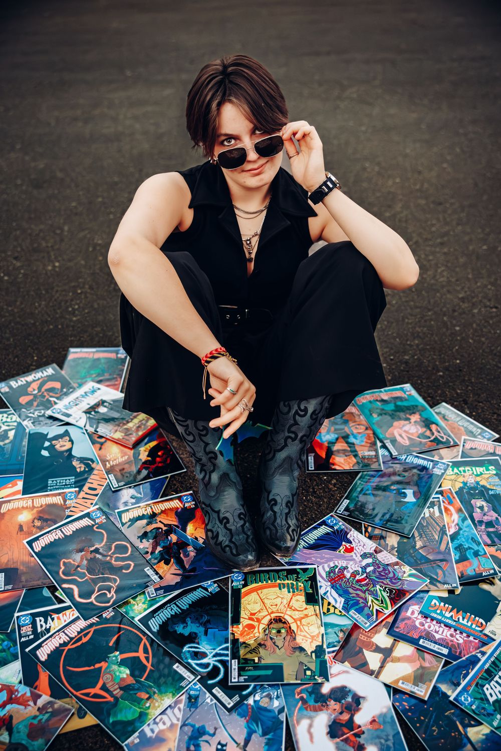 Young woman in sunglasses sits on pavement surrounded by colorful comic books in black outfit.