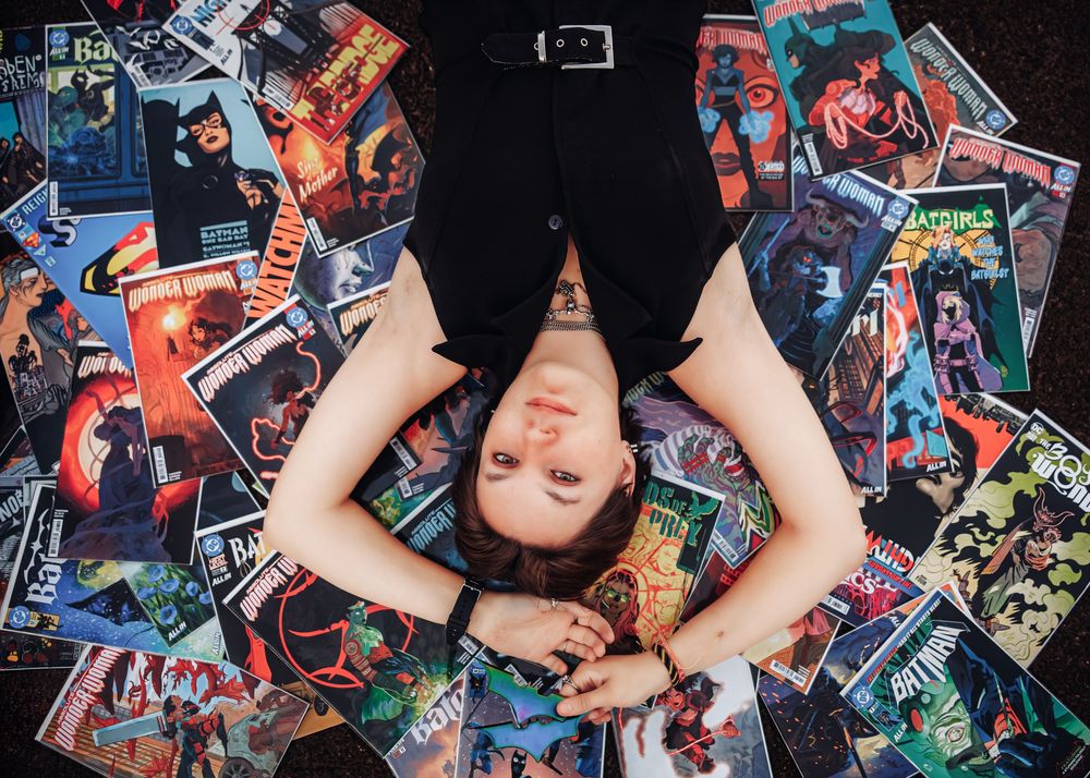 Person lying among colorful Batman comic books, viewed from above, wearing black outfit.