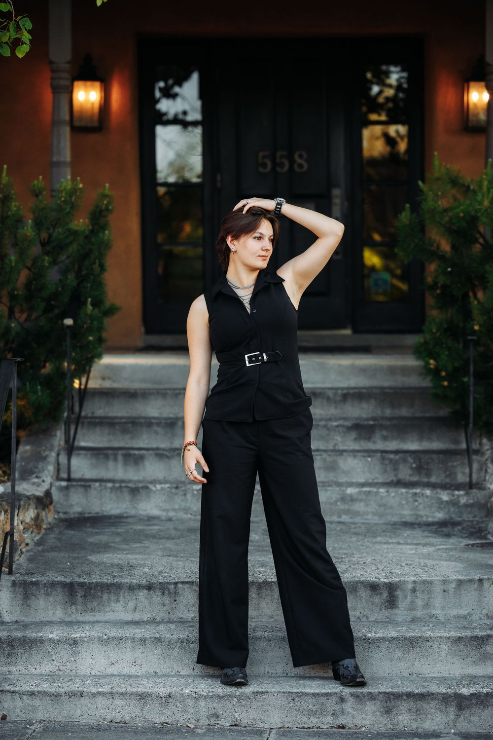 Woman in black sleeveless jumpsuit with belt poses on stone steps outside building number 558.