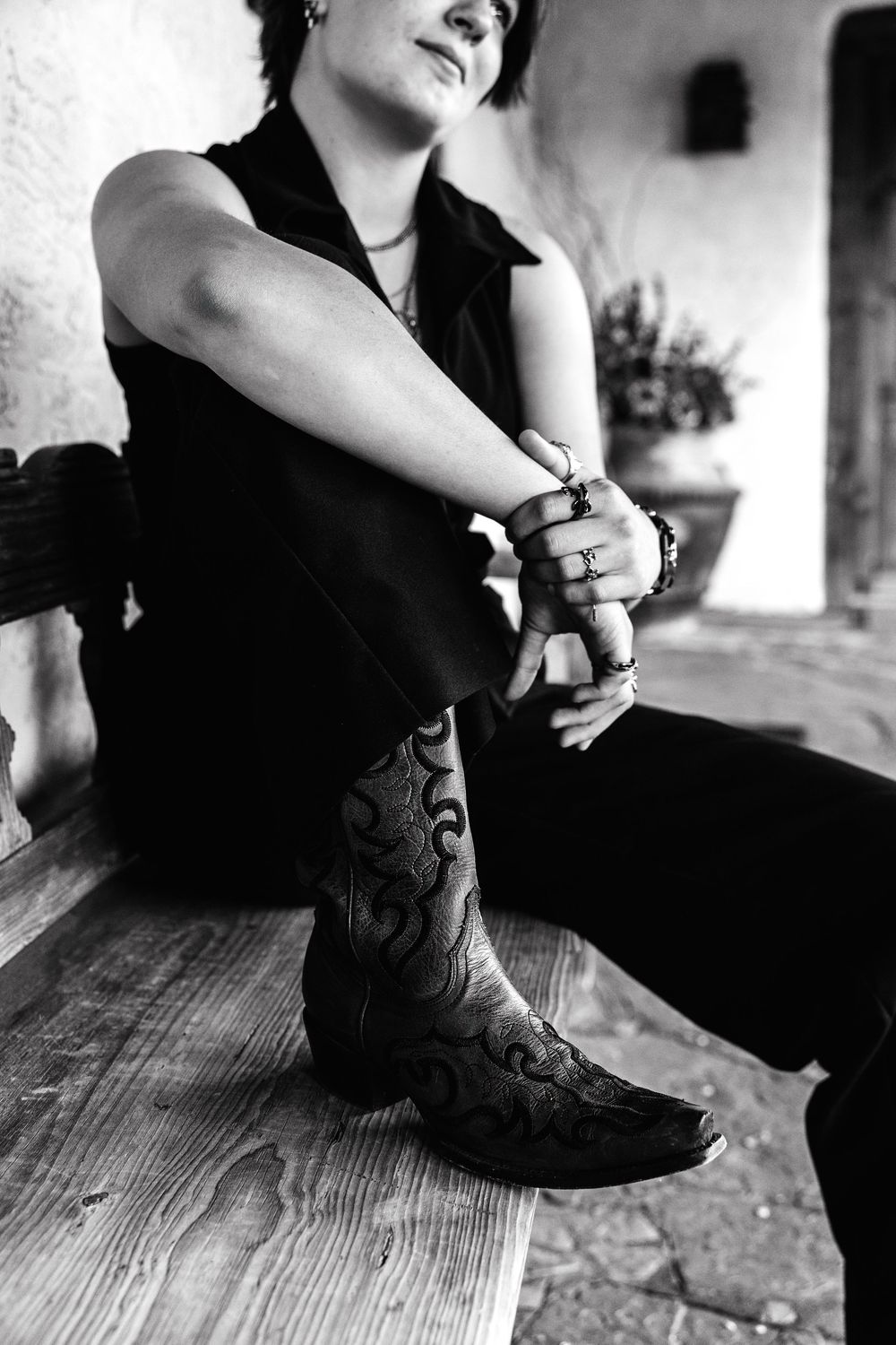 Black and white photo of a person sitting on wooden steps wearing ornate cowboy boots, rings, and bracelets.