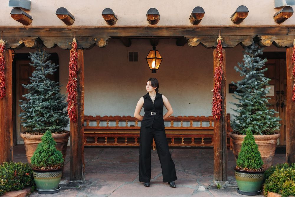 Woman in black jumpsuit posing in front of a Southwestern adobe building with red chili ristras and pine trees.