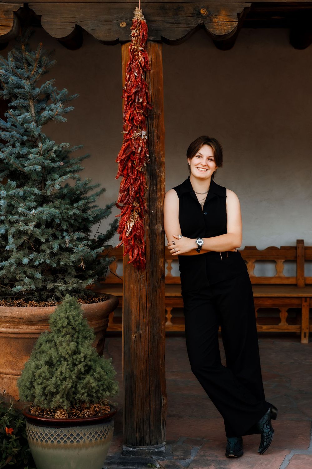 Woman in black outfit smiling beside a wooden post with red chili ristras, surrounded by potted evergreen trees.