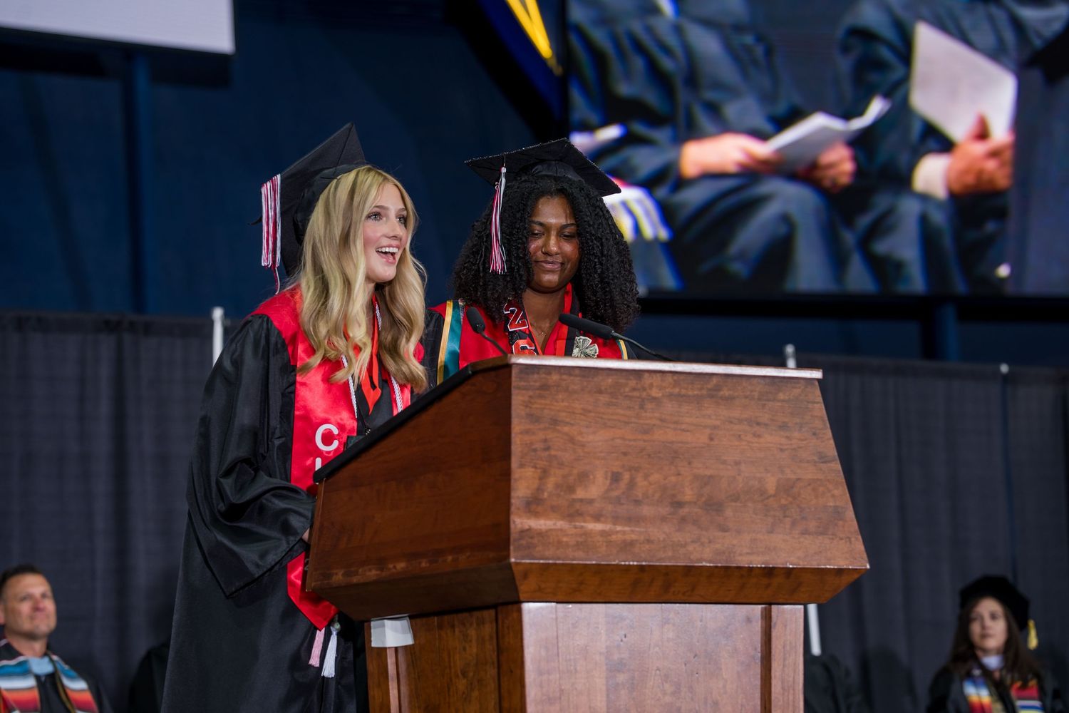 Two graduates in black caps and gowns speak at a podium during a college graduation ceremony.