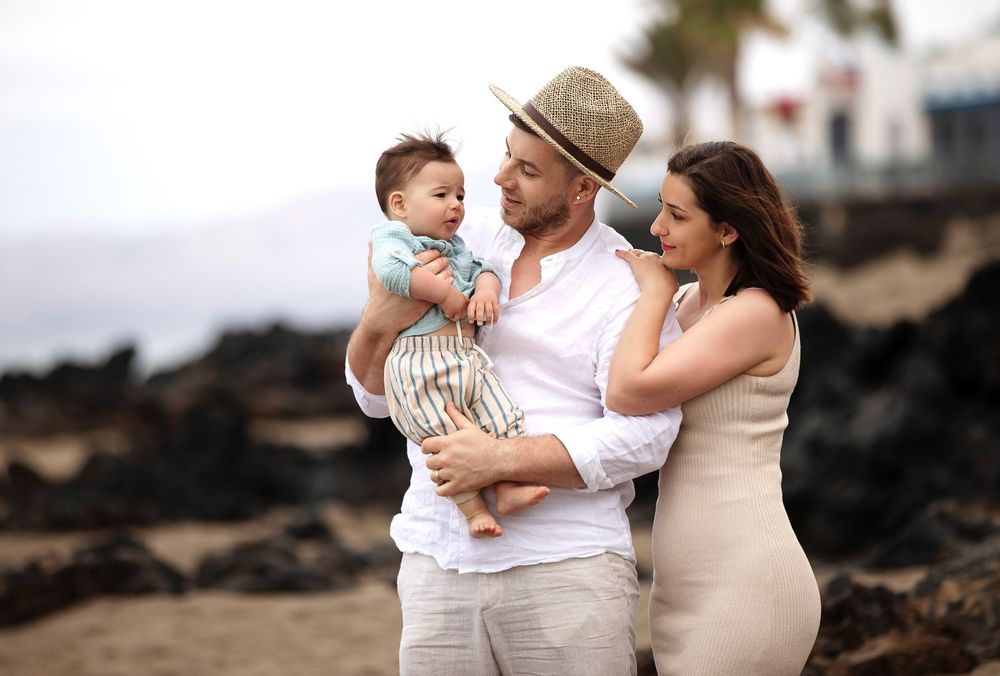 A couple holding their baby boy on a rocky beach, dressed in light summer outfits, sharing a tender family moment.
