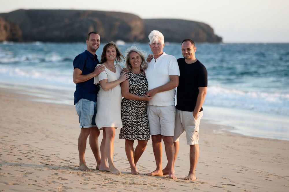 Family of five smiling together on a sandy beach at sunset with ocean waves and rocky cliffs in the background.