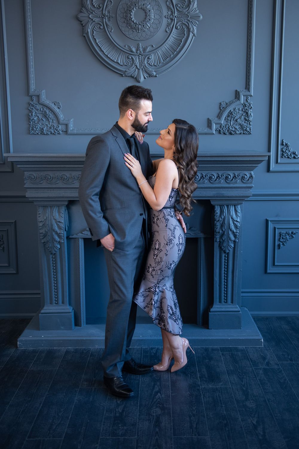 Couple in formal attire sharing an intimate moment by an ornate gray fireplace.