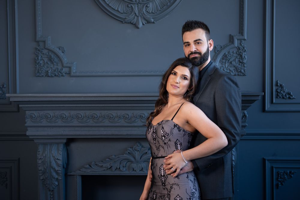 Elegant couple posing together in formal wear before a decorative gray fireplace.