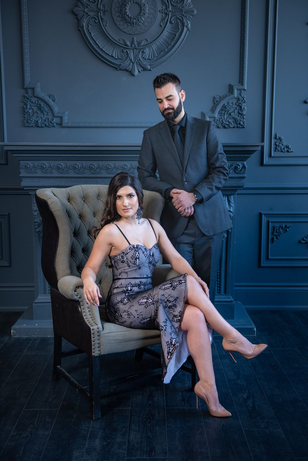 Stylish couple in formal attire posing in an ornate dark room, woman seated in vintage armchair.