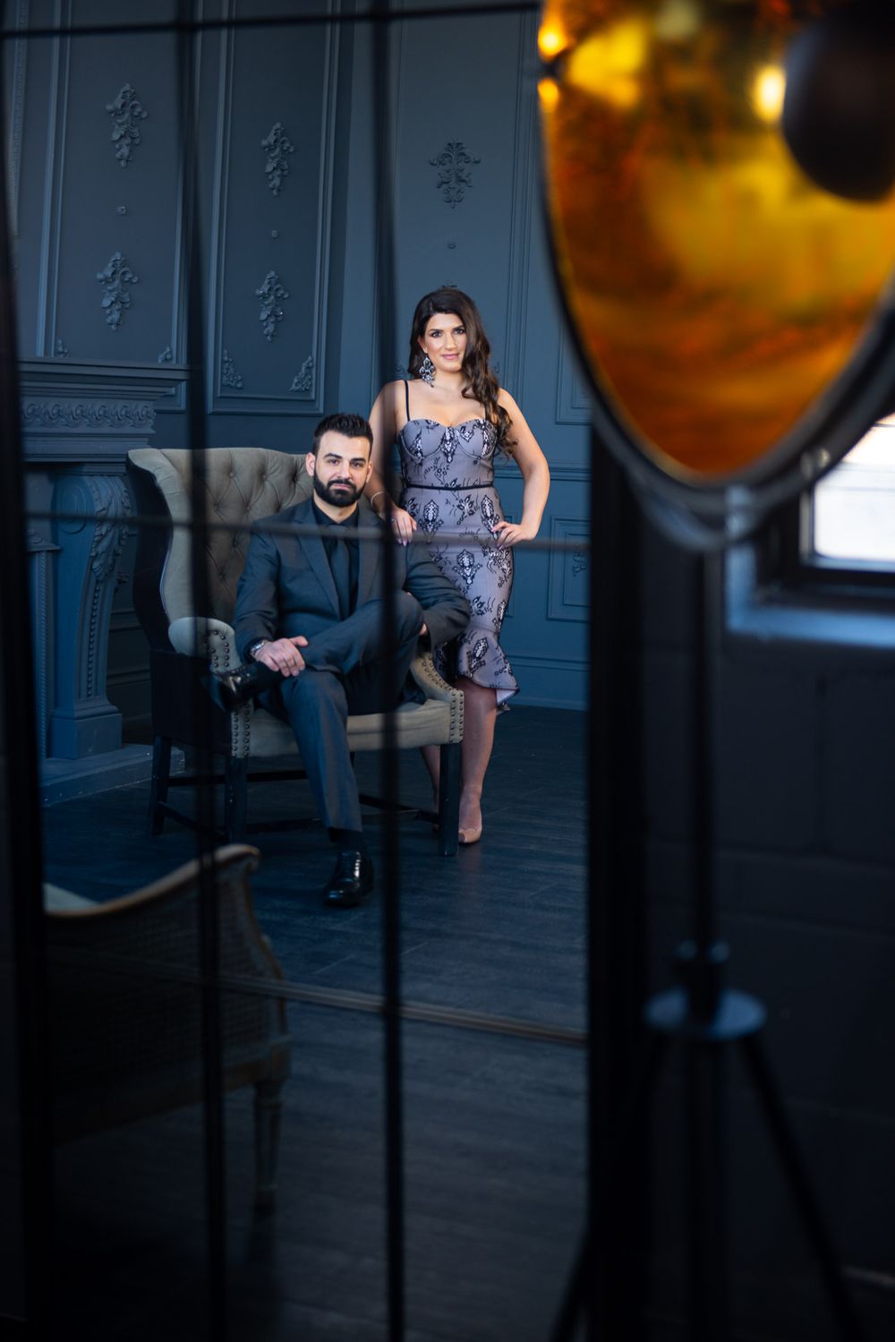Sophisticated couple in elegant formalwear posing in a dark luxury interior with ornate moldings.