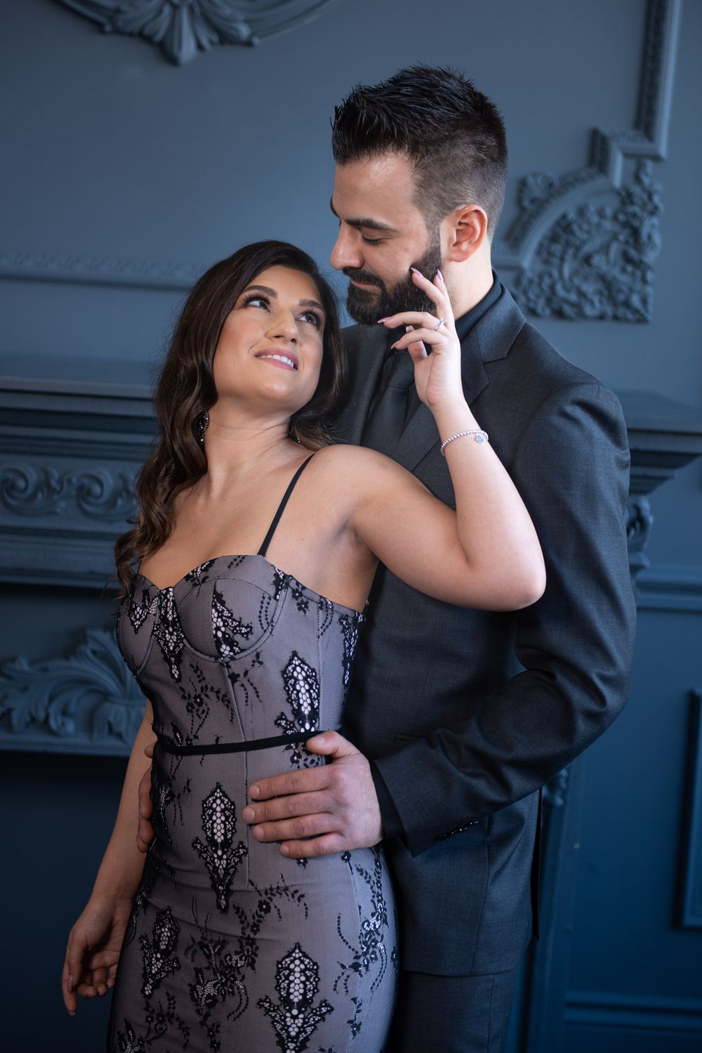 An elegant couple embraces warmly, smiling together against a classic blue-gray wall.
