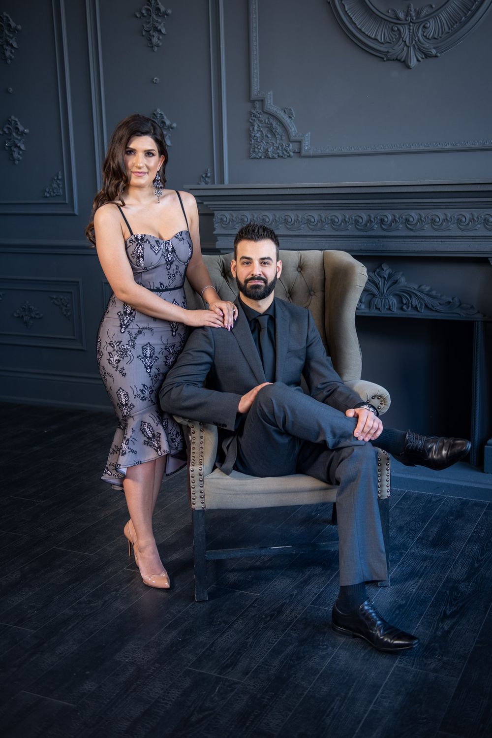 Elegant couple posing in luxury gray interior, man seated in vintage chair, woman in lace dress beside him.