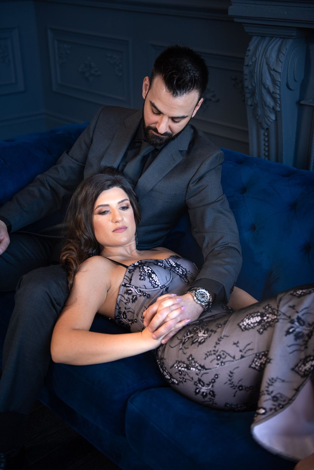 Romantic couple on blue velvet sofa, woman in lace bodysuit reclining against bearded man in dark suit.