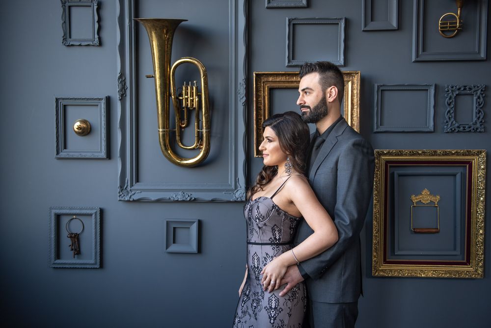 Stylish couple embracing before decorative wall with gold frames and brass tuba in moody blue setting.