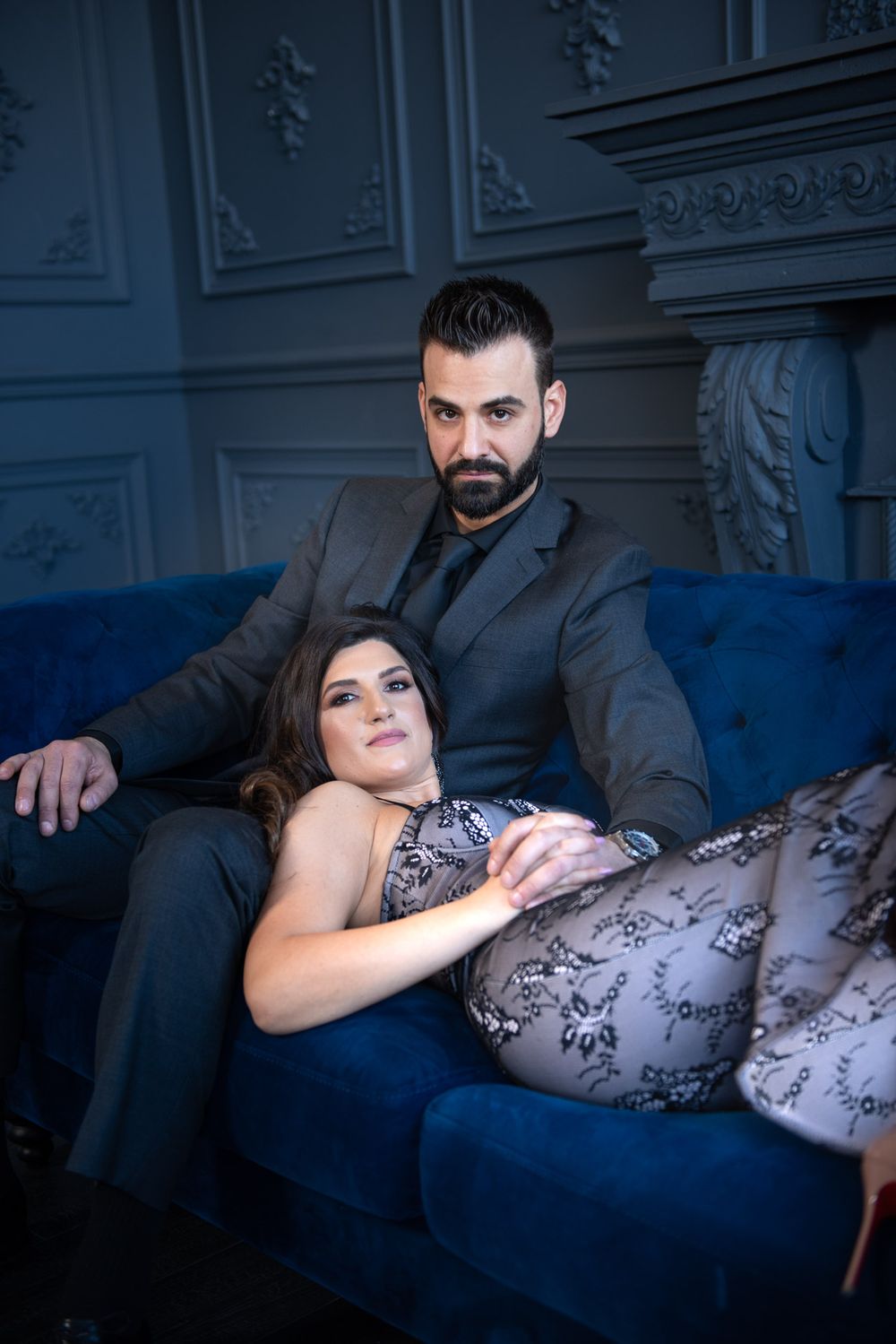 Couple posing on blue velvet couch in elegant dark room, woman reclining across mans lap looking at camera.