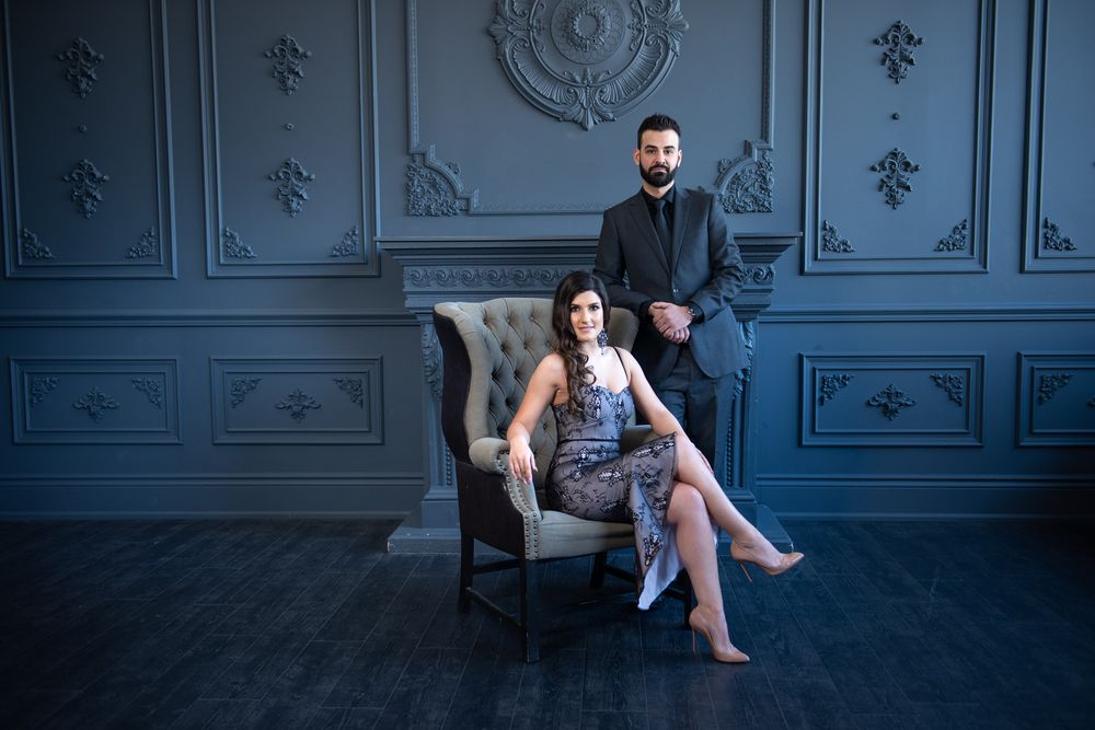 Couple in elegant gray room, woman seated in vintage chair wearing lace dress, man standing behind her.