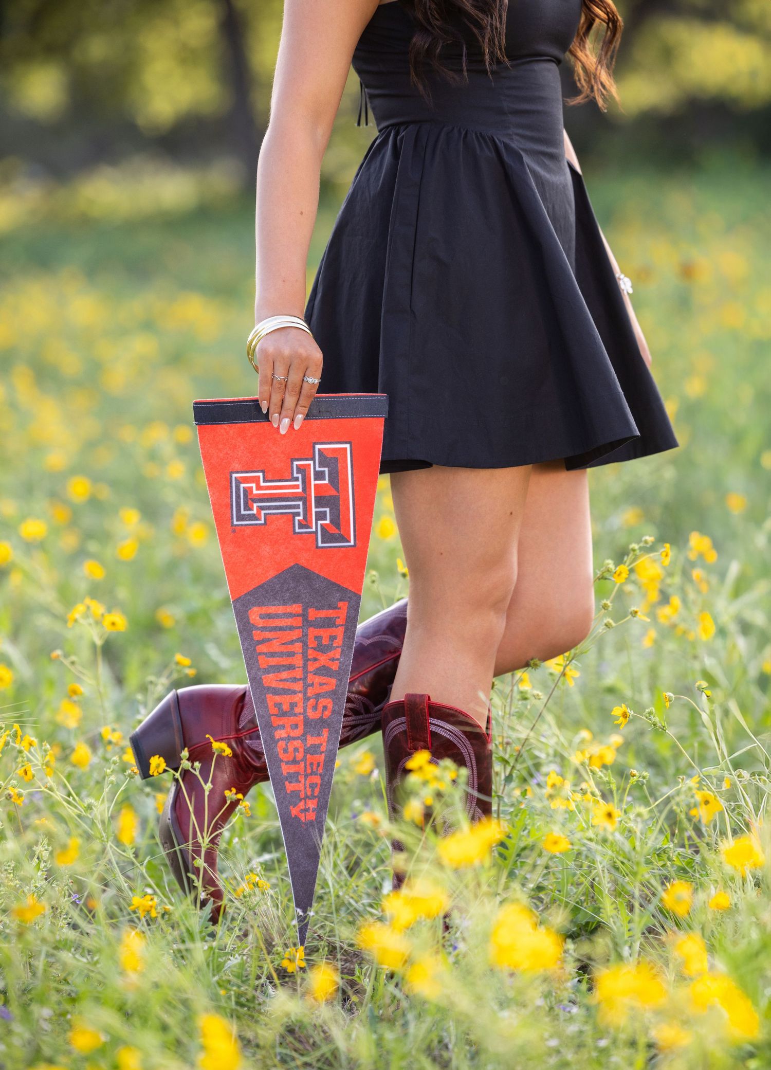 2027 senior girl in black dress and cowboy boots holding a Texas Tech University pennant in a field of yellow wildflowers.