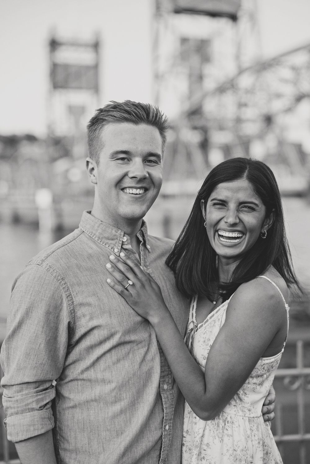 Black and white portrait of a laughing couple posing together near a lift bridge.