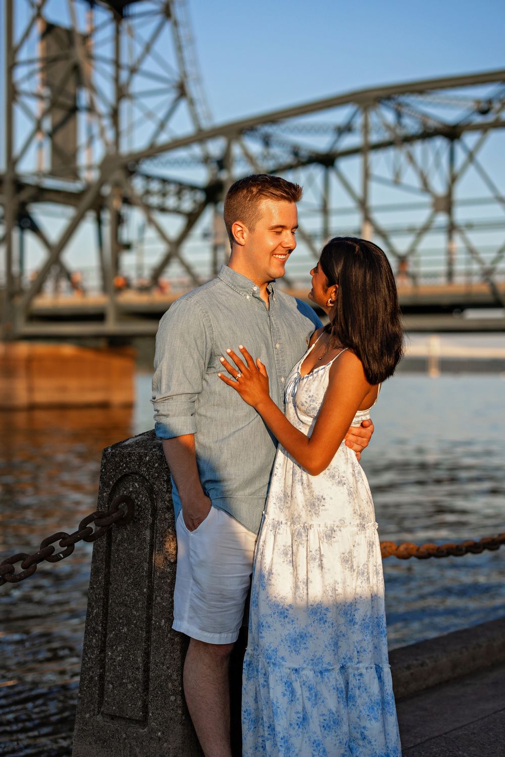 Engaged couple smiling at each other on a waterfront dock with a lift bridge behind them.