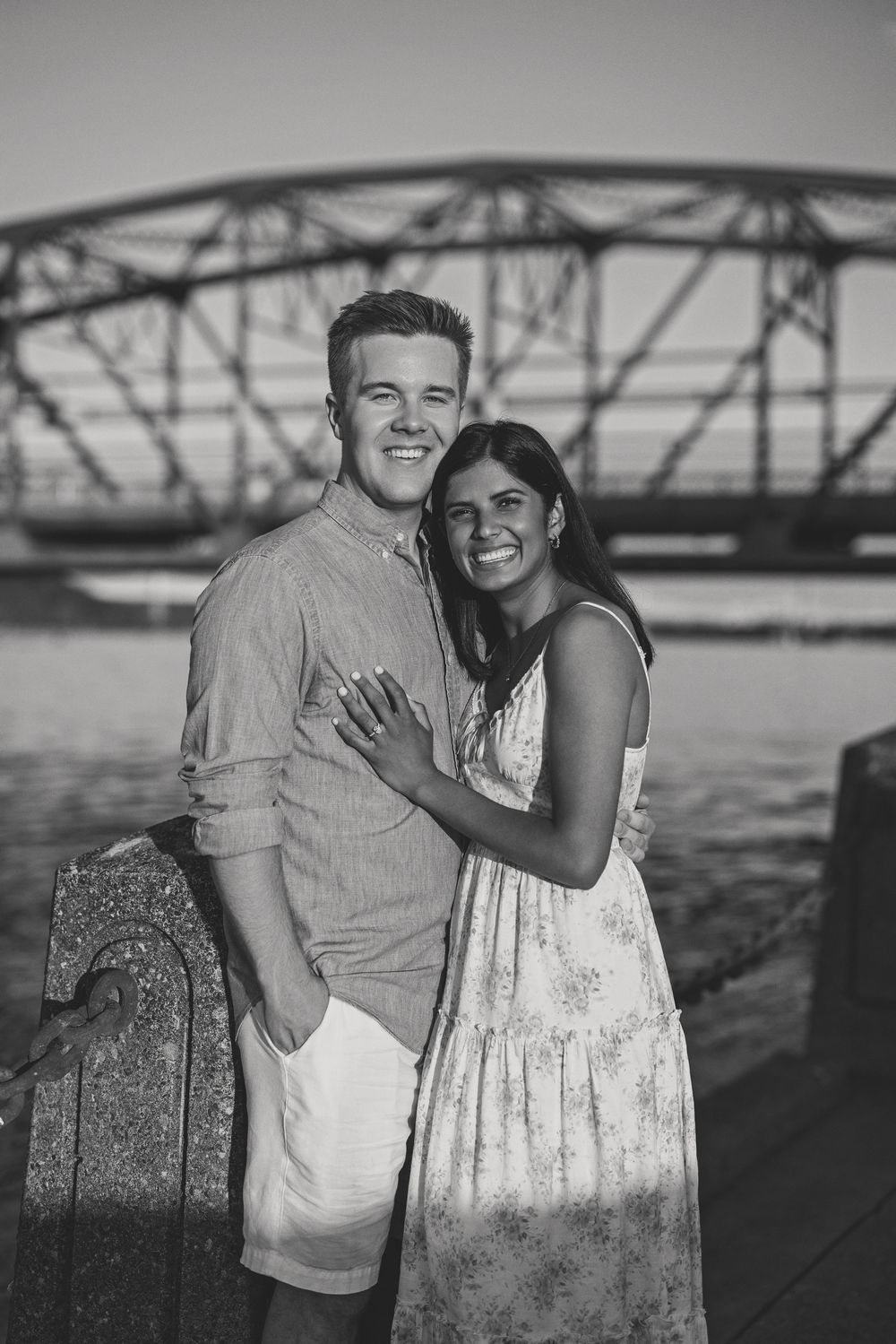 Black and white photo of a smiling couple embracing on a dock with a bridge backdrop.