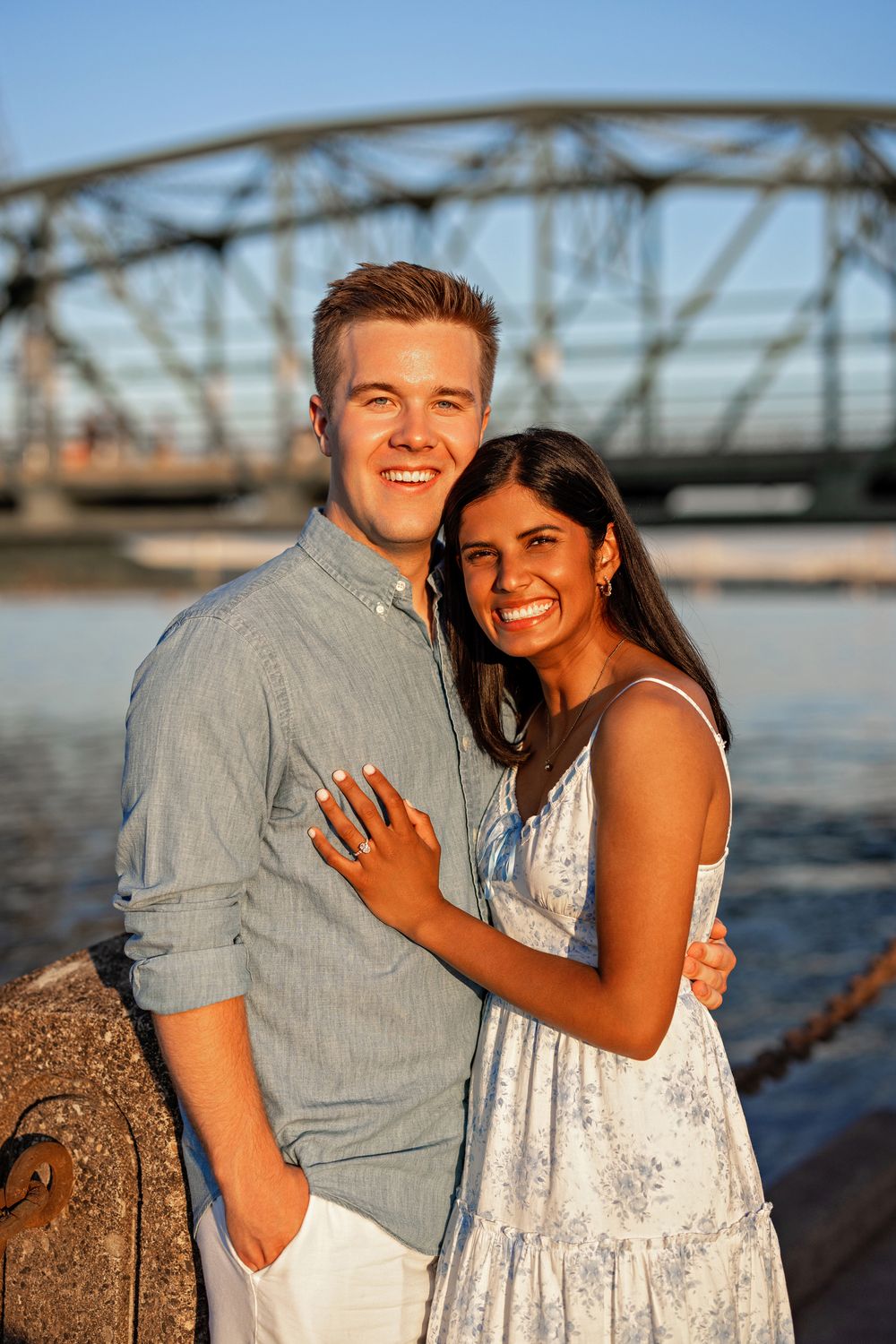 Smiling couple posing together on a sunny waterfront with a steel lift bridge behind them.