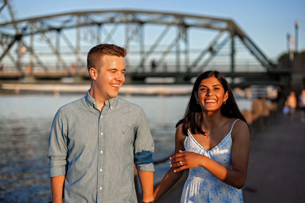 Happy couple smiling and walking hand-in-hand along a waterfront promenade with a steel bridge in the background.