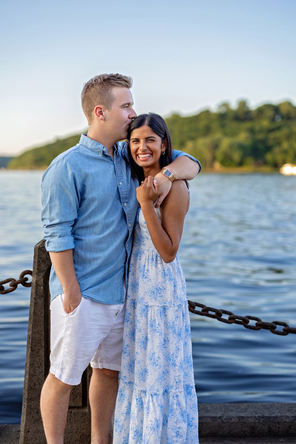 Loving couple embracing on a waterfront dock, man in blue shirt and woman in floral dress, green hills behind.