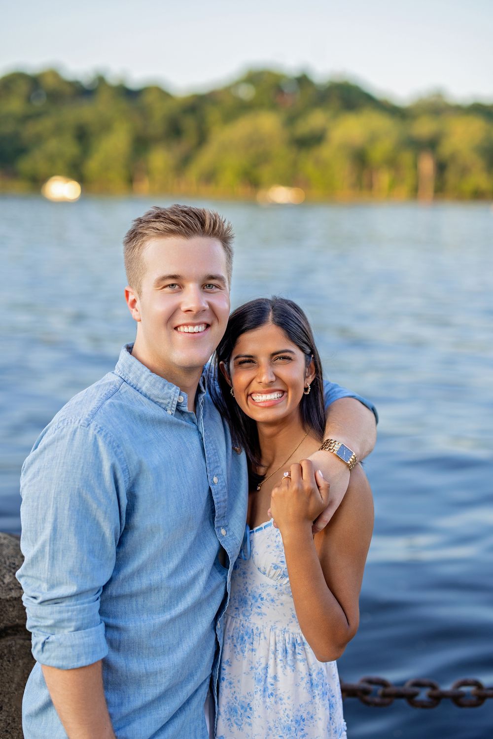 Smiling couple posing together by a waterfront chain fence, woman in blue floral dress laughing joyfully.