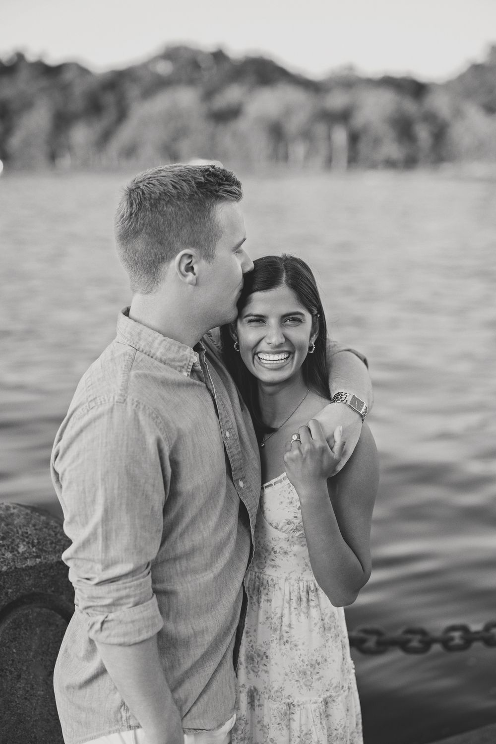 Black and white photo of a couple laughing together by the water, man whispering in womans ear.