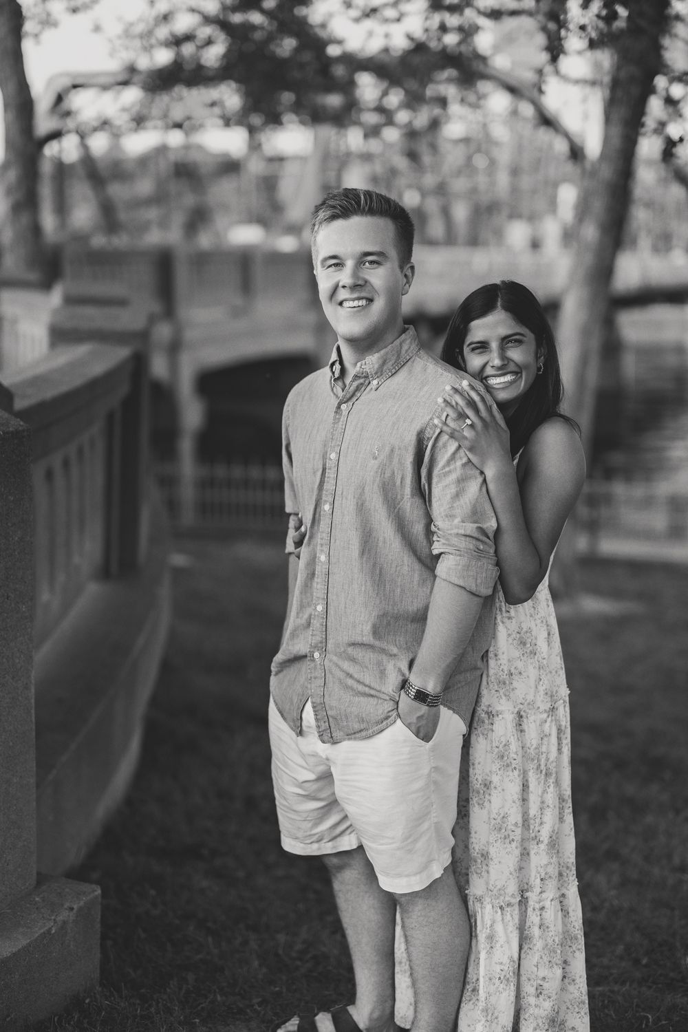 Black and white photo of a smiling couple outdoors, woman hugging man from behind near a stone bridge.